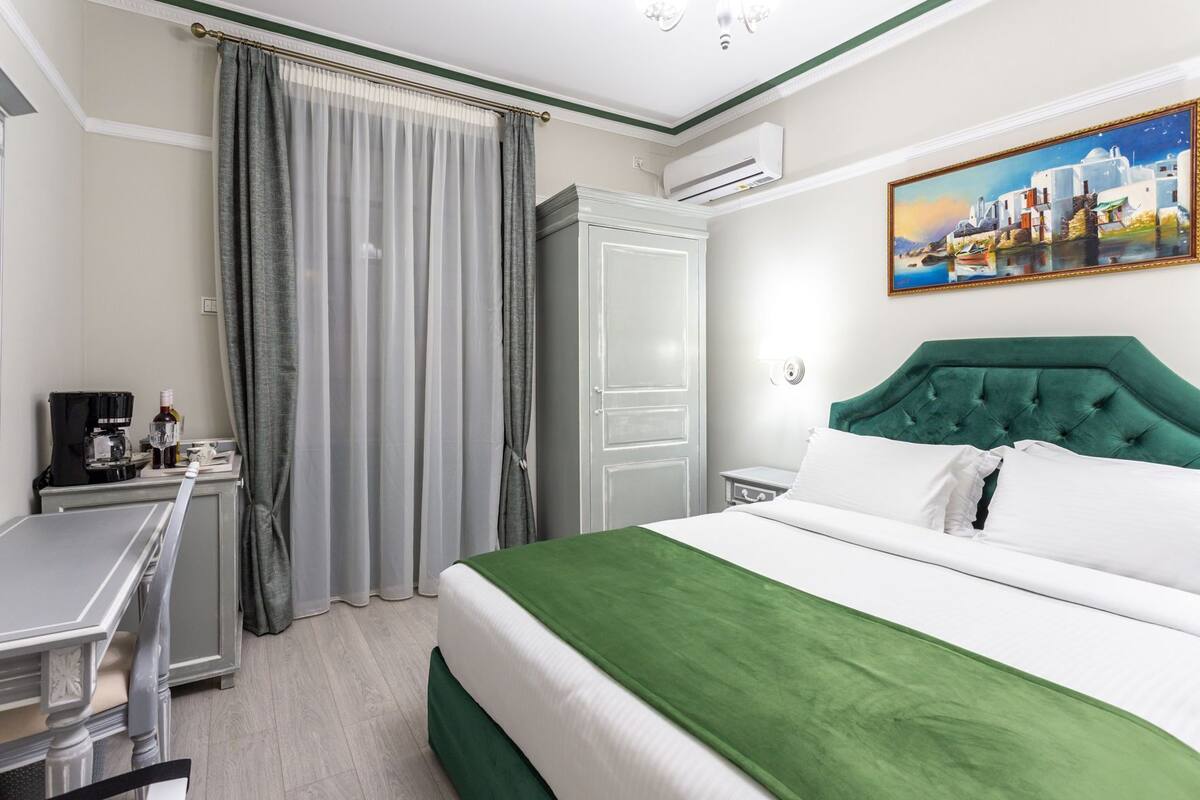 A hotel room with a bed, a desk, a chair, a wardrobe, and a painting on the wall.