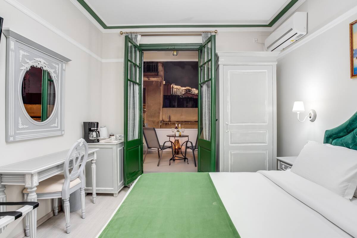 A hotel room with a green bedspread, a desk with a chair, a dining table with chairs, and a view of the outside balcony.