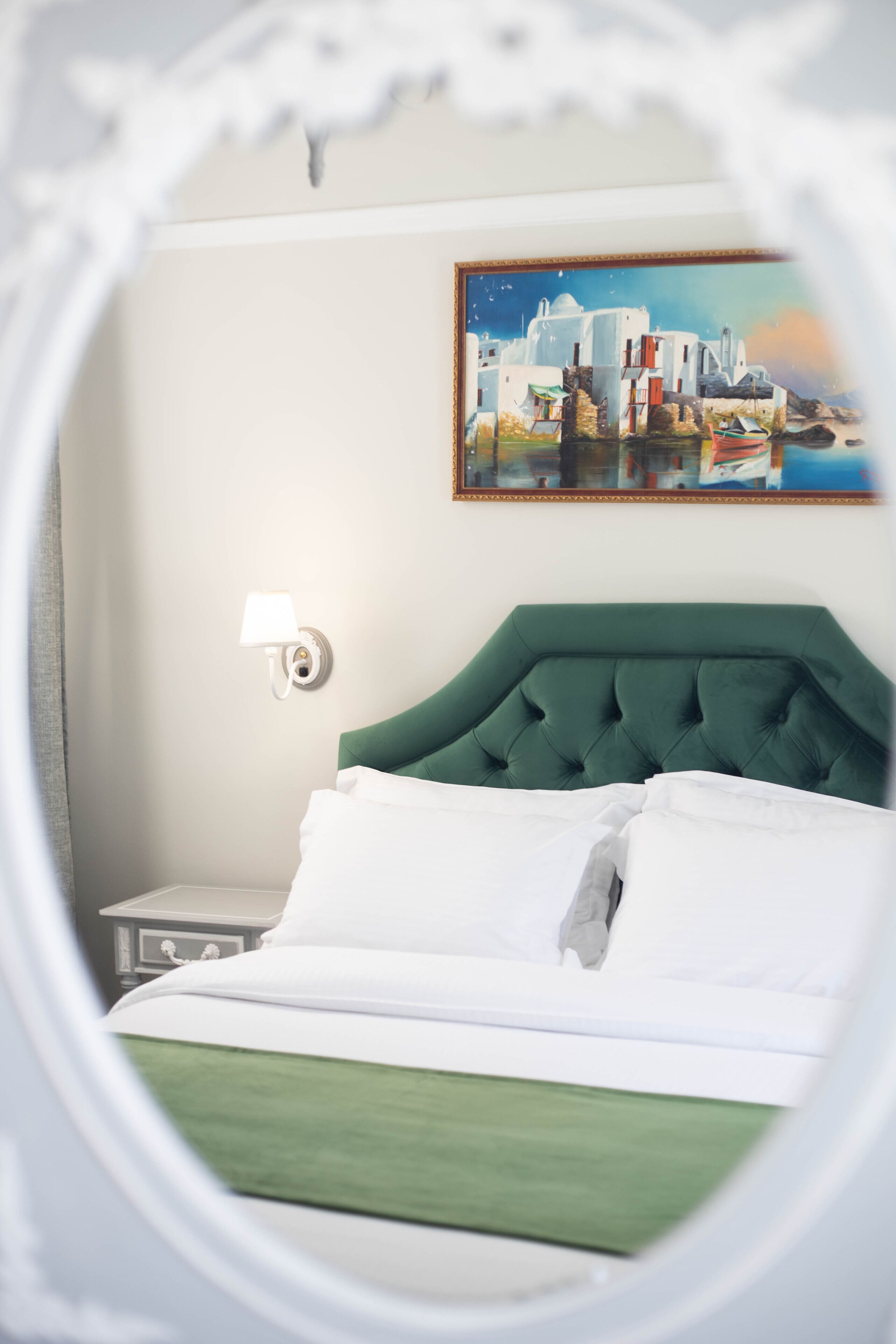 A hotel room with a green upholstered headboard, white bedding, a painting of a coastal scene, and a bedside lamp.