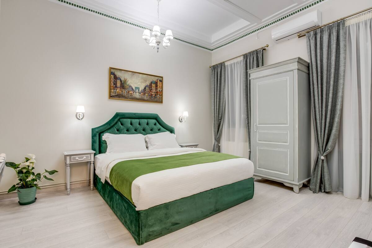 A neatly arranged bedroom with a green upholstered bed, a white nightstand, a wall-mounted painting, and a large wardrobe.