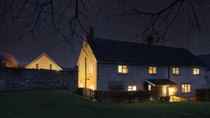 Una casa a due piani con un muro in pietra e un tetto spiovente, illuminata da luci interne su uno sfondo di cielo notturno.