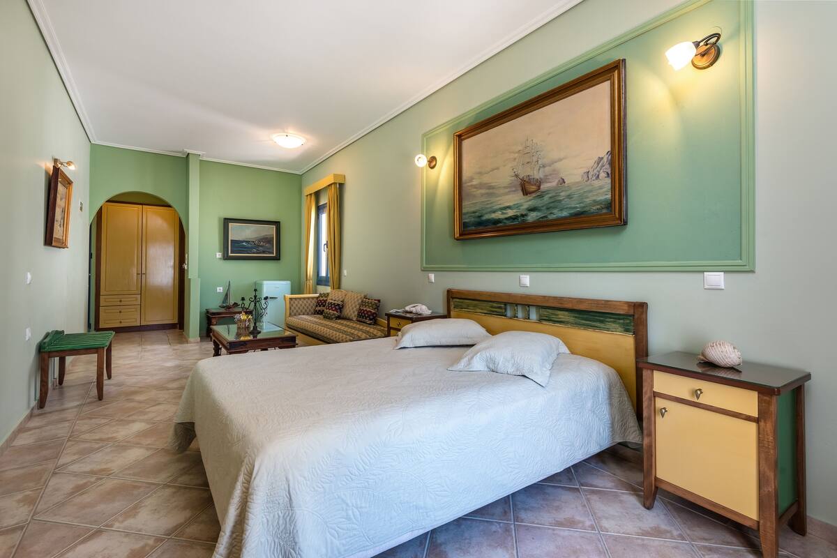 Une chambre à coucher avec un grand lit, une table de chevet, une commode, un fauteuil vert et un tableau au mur.