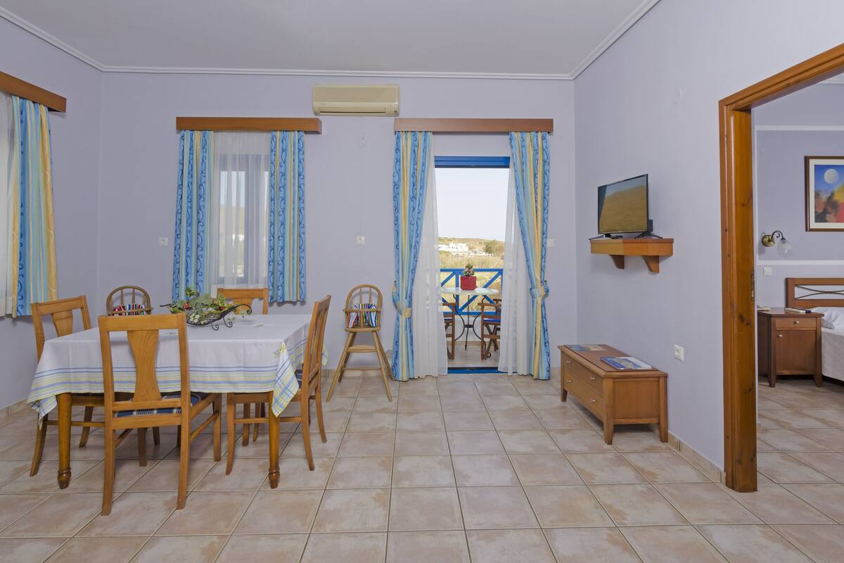 Un coin repas avec une table dressée pour quatre personnes, des chaises en bois, une petite table d’appoint et un téléviseur posé sur une étagère.