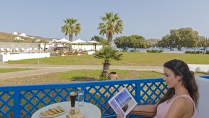 Une femme est assise à une table, devant une boisson et de la nourriture, et elle jouit d’une vue panoramique sur un paysage agrémenté de palmiers et d’une étendue d’eau.
