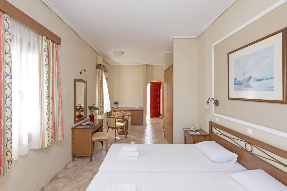Une chambre d’hôtel avec deux lits, un bureau, une chaise, une commode et une grande fenêtre avec des rideaux.