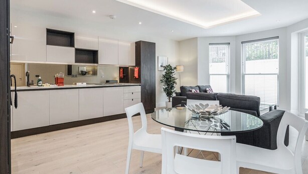 A modern kitchen with white cabinets, a dining area with a glass table and white chairs, and a living area with a dark sofa and a potted plant.