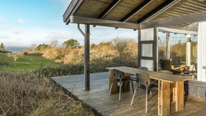 Eine Holzterrasse mit Tisch und Stühlen, die auf eine grüne Fläche mit Bäumen und Meerblick blickt.