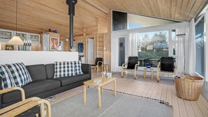 Ein gemütliches Wohnzimmer mit einer Couch, Sesseln, einem Couchtisch und einem großen Fenster mit Blick ins Freie.