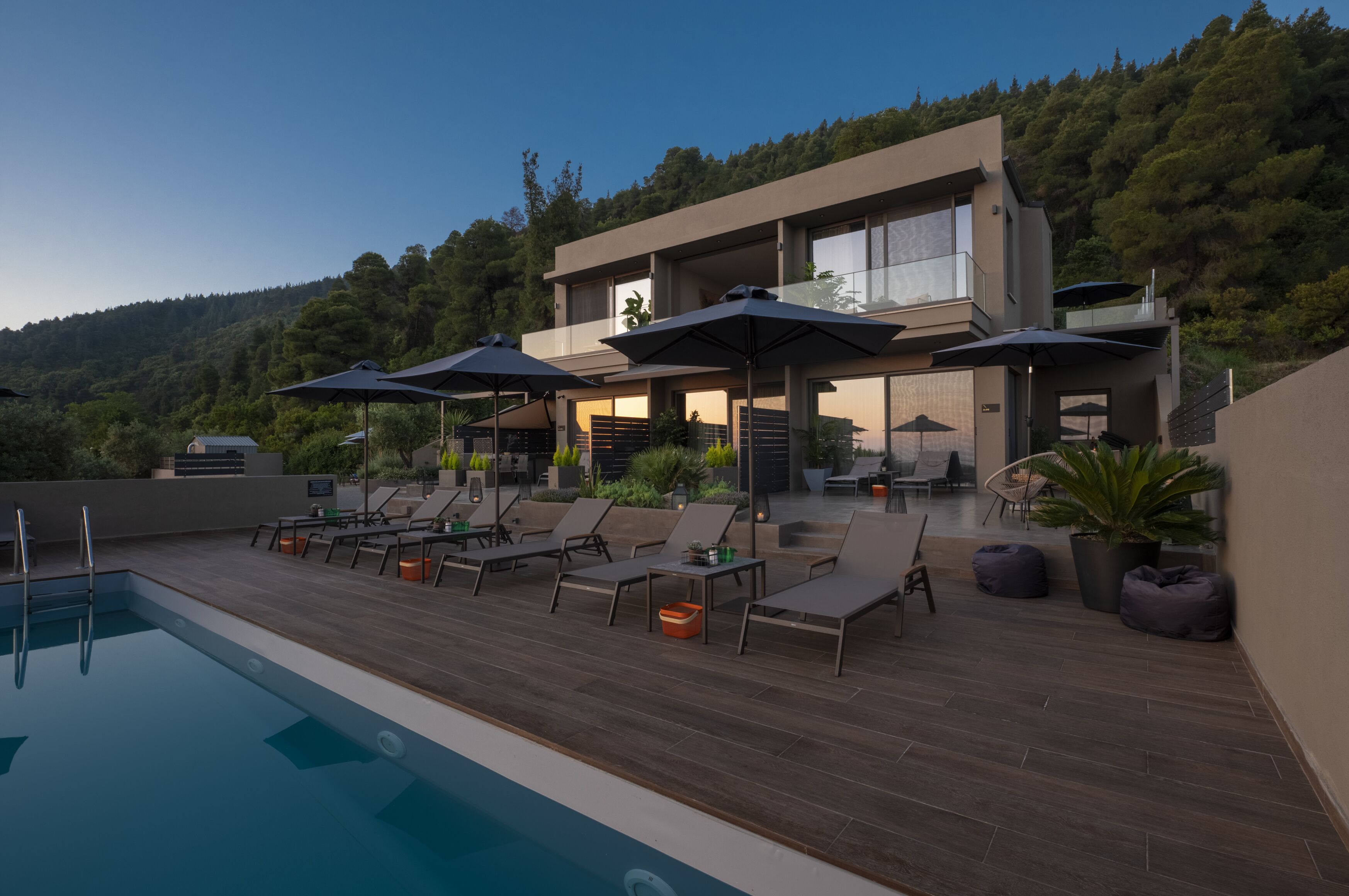 A modern outdoor pool area with lounge chairs and umbrellas, surrounded by a wooden deck and potted plants.
