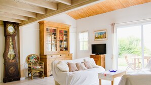 A cozy living room with a wooden ceiling, a large wooden cabinet, a television, a covered sofa, and a view of the outdoors through a large window. - House for 6 in the heart of Saint Martin (Saint-Martin-de-Ré)