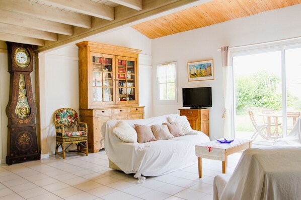A cozy living room with a wooden ceiling, a large wooden cabinet, a television, a covered sofa, and a view of the outdoors through a large window. - House for 6 in the heart of Saint Martin (Saint-Martin-de-Ré)