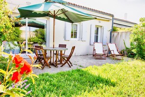 A patio with a round table and chairs, a green umbrella, and a white building with windows and shutters. - House for 6 in the heart of Saint Martin (Saint-Martin-de-Ré)