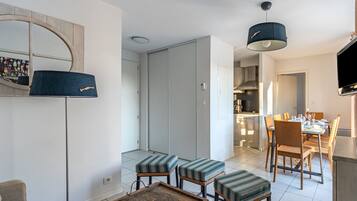 Un salon moderne comprenant un canapé, des poufs, une table basse, un coin repas avec des chaises et une table, une cuisine en arrière-plan et un téléviseur fixé au mur.