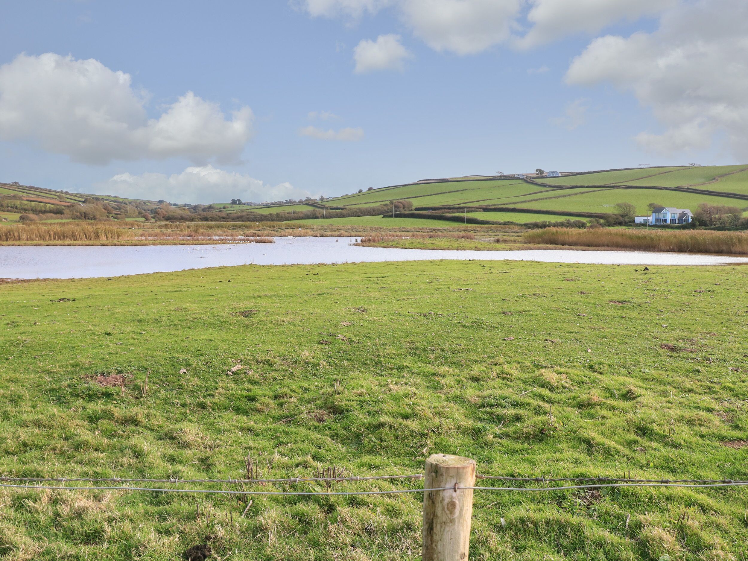 A rural landscape with a pond, grassy fields, and a house in the distance.