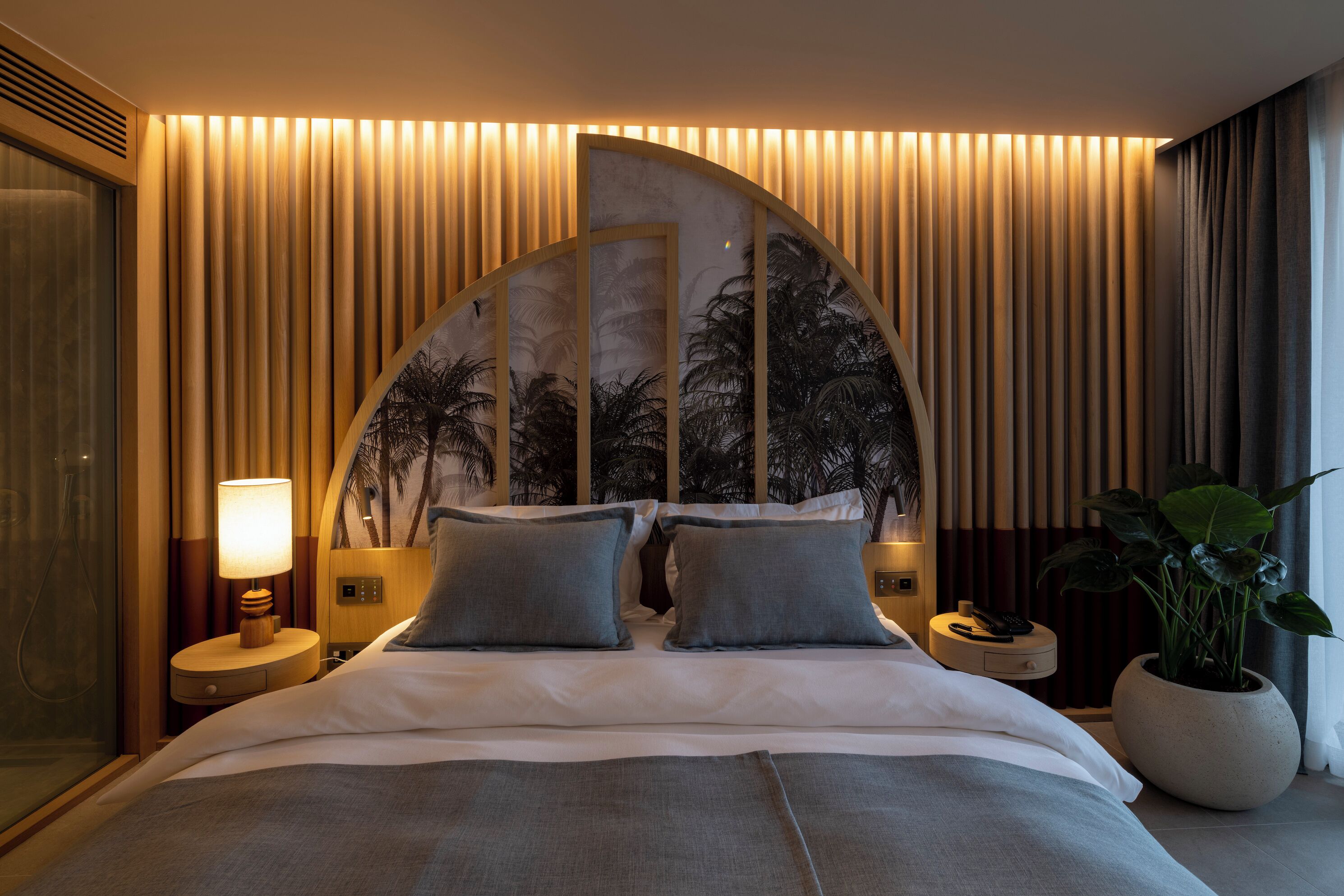 A hotel room with a curved headboard, a bed with grey bedding, a nightstand with a lamp, a potted plant, and a view of palm trees through the headboard.