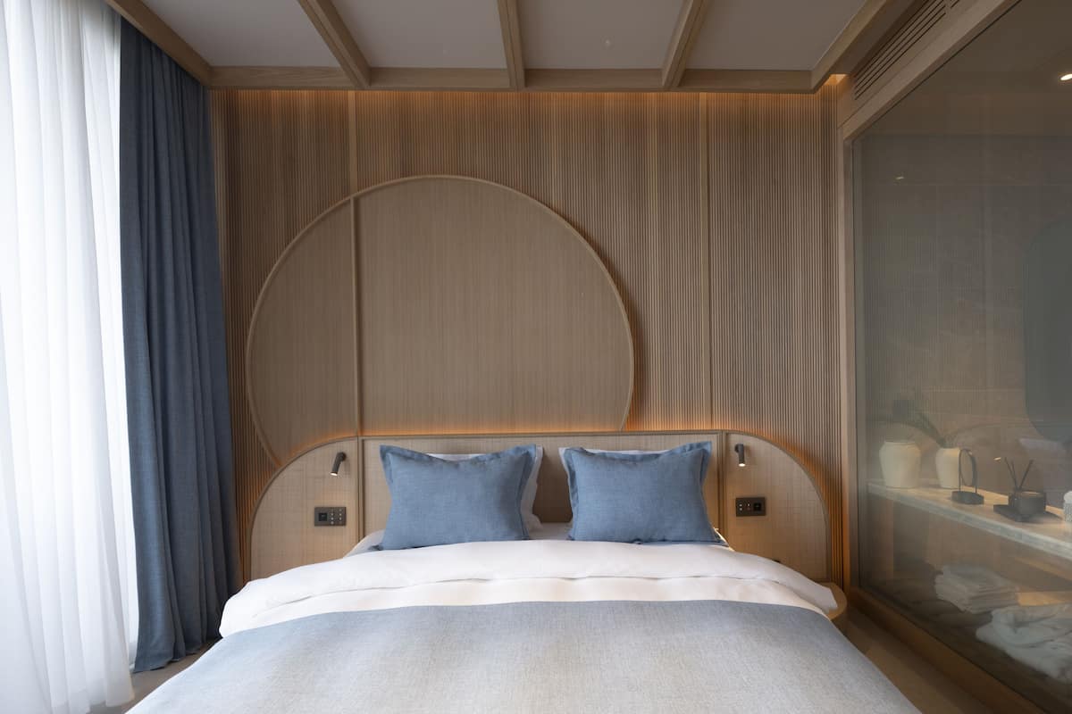 A modern hotel room with a large bed, a wooden headboard, and a view of the city through a large window.