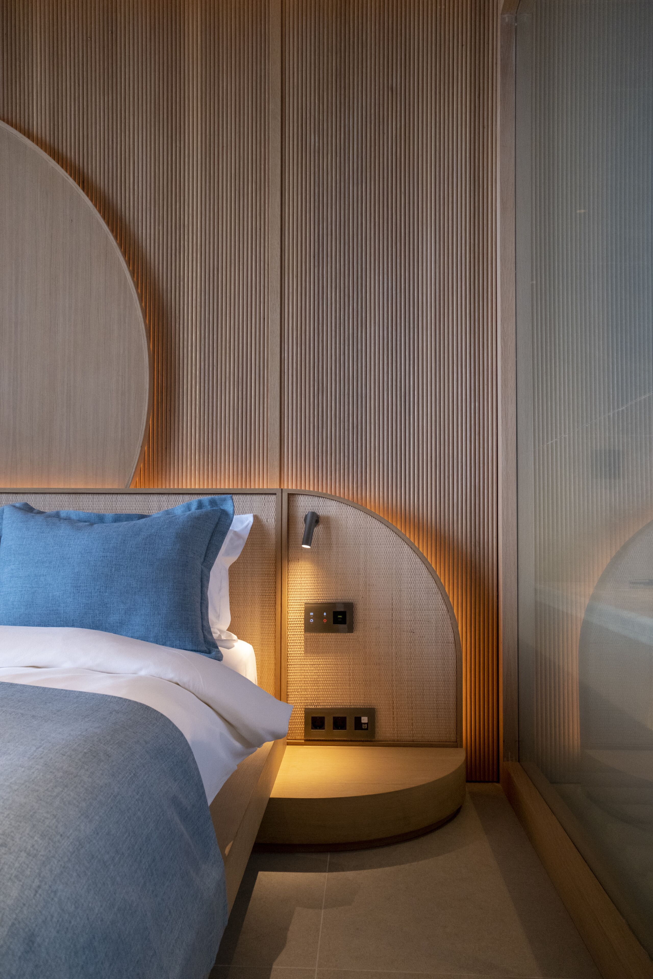 A modern bedroom with a curved headboard, a bed with blue bedding, and a small bedside table with a lamp.