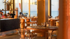 A restaurant with tables and chairs, a bar area, and large windows offering a view of the sea.