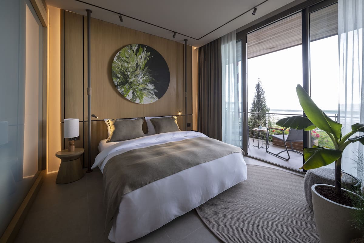 A modern hotel room with a large bed, a round painting on the wall, a potted plant, and a balcony with a view of the sea.