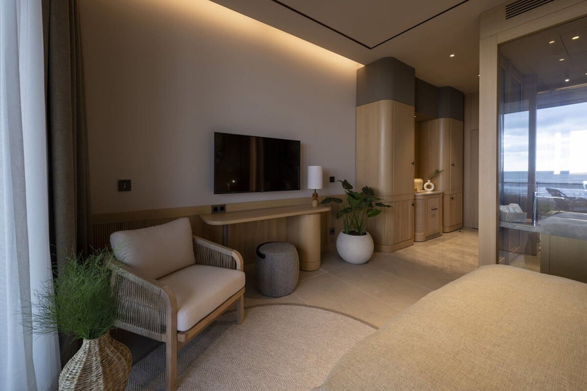 A modern hotel room with a flat-screen TV, a sofa, a potted plant, and a view of the sea.
