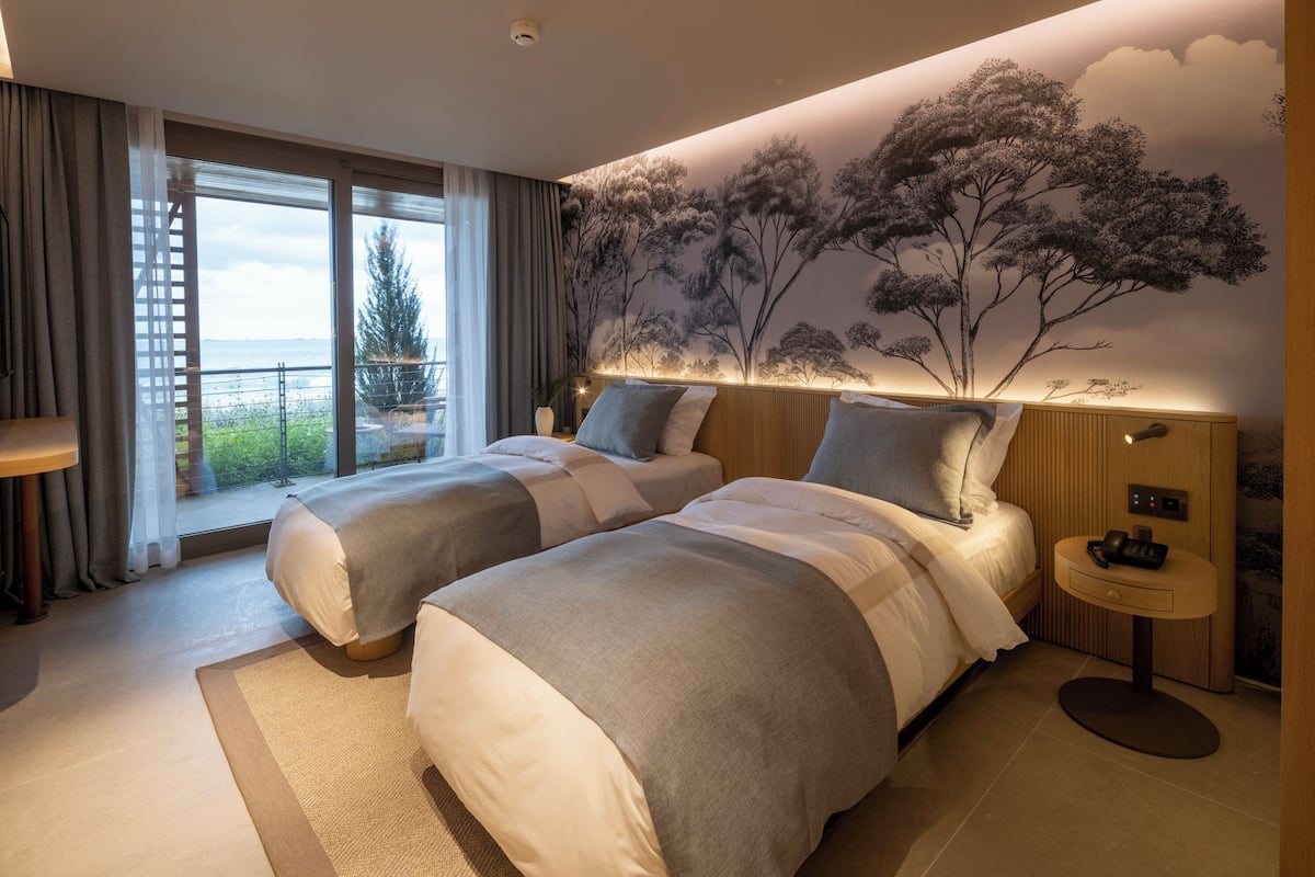 A hotel room with two beds, a large window, a mural of trees, and a small table with a phone.