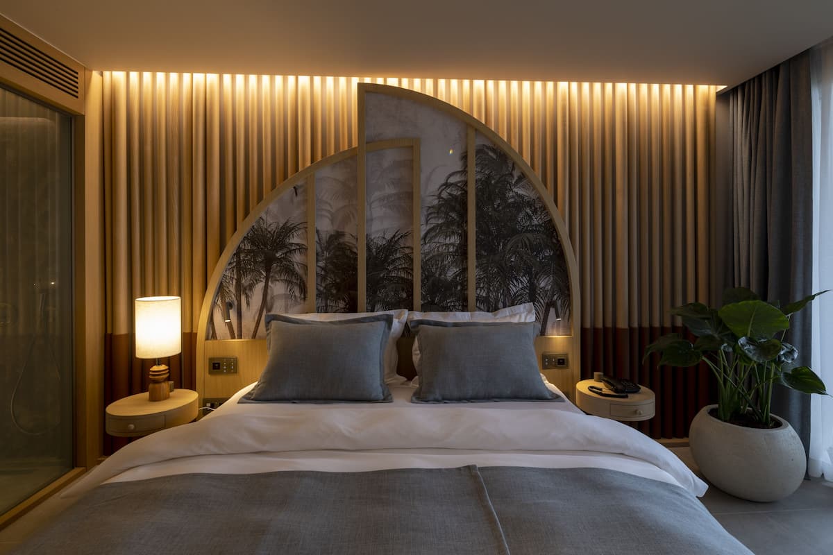 A modern hotel room with a curved headboard, a large bed, a bedside table with a lamp, a nightstand, and a potted plant.