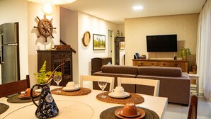 A dining area with a table set for two, a television in the background, and a sofa in the living room. - MSP02 Village 3 Quartos Pé na Areia (Cairu)