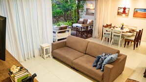 A living room with a sofa, dining table, chairs, and a view of a garden. - MSP02 Village 3 Quartos Pé na Areia (Cairu)