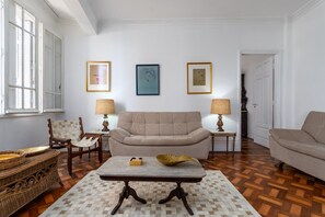 A living room with a beige sofa, two armchairs, a coffee table, and a patterned rug. There are three framed artworks on the wall and two lamps on side tables.
