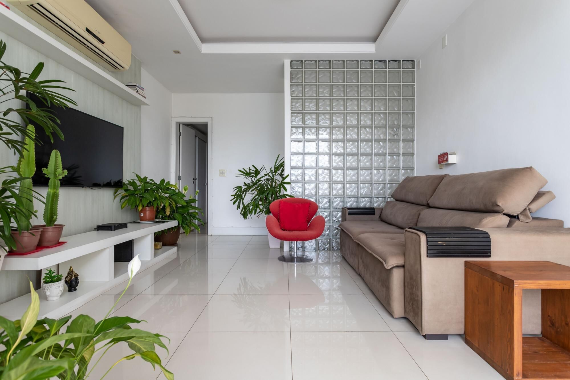 A modern living room with a beige sofa, a red chair, and various potted plants.