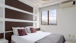 A bedroom with a large bed, a view of the city through a window, and an air conditioning unit on the wall. - Coverage in Barra da Tijuca for 6 People Lc2207 (Rio de Janeiro)