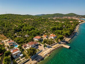 Luftaufnahme einer Küstenlandschaft mit Häusern, tiefblauem Meer und grünen Hügeln im Hintergrund.