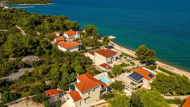 An aerial view of a coastal residential area with houses featuring red-tiled roofs, surrounded by lush greenery and a clear blue sea. - Villa Tatijana Zadarvillas (Kukljica)
