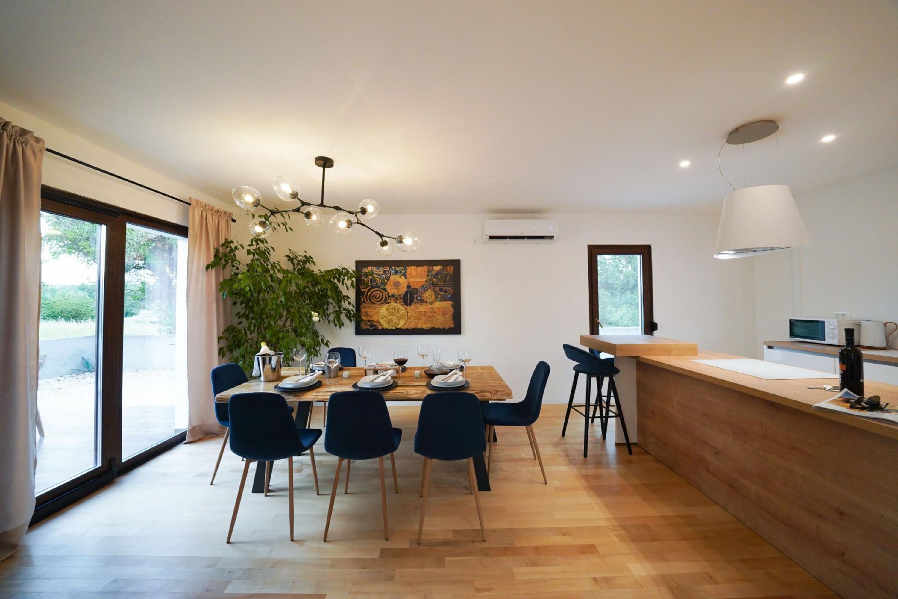 A modern dining area with a wooden table, blue chairs, a hanging light fixture, and a large window.