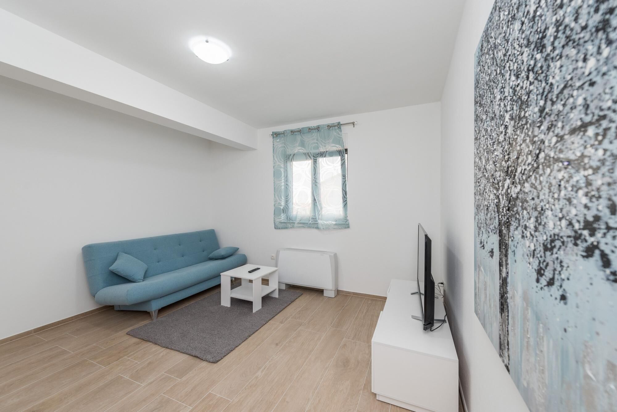 Ein modernes Wohnzimmer mit einem blauen Sofa, einem weißen Couchtisch, einem kleinen Teppich, einer Heizung und einem Fernseher auf einem Lowboard.