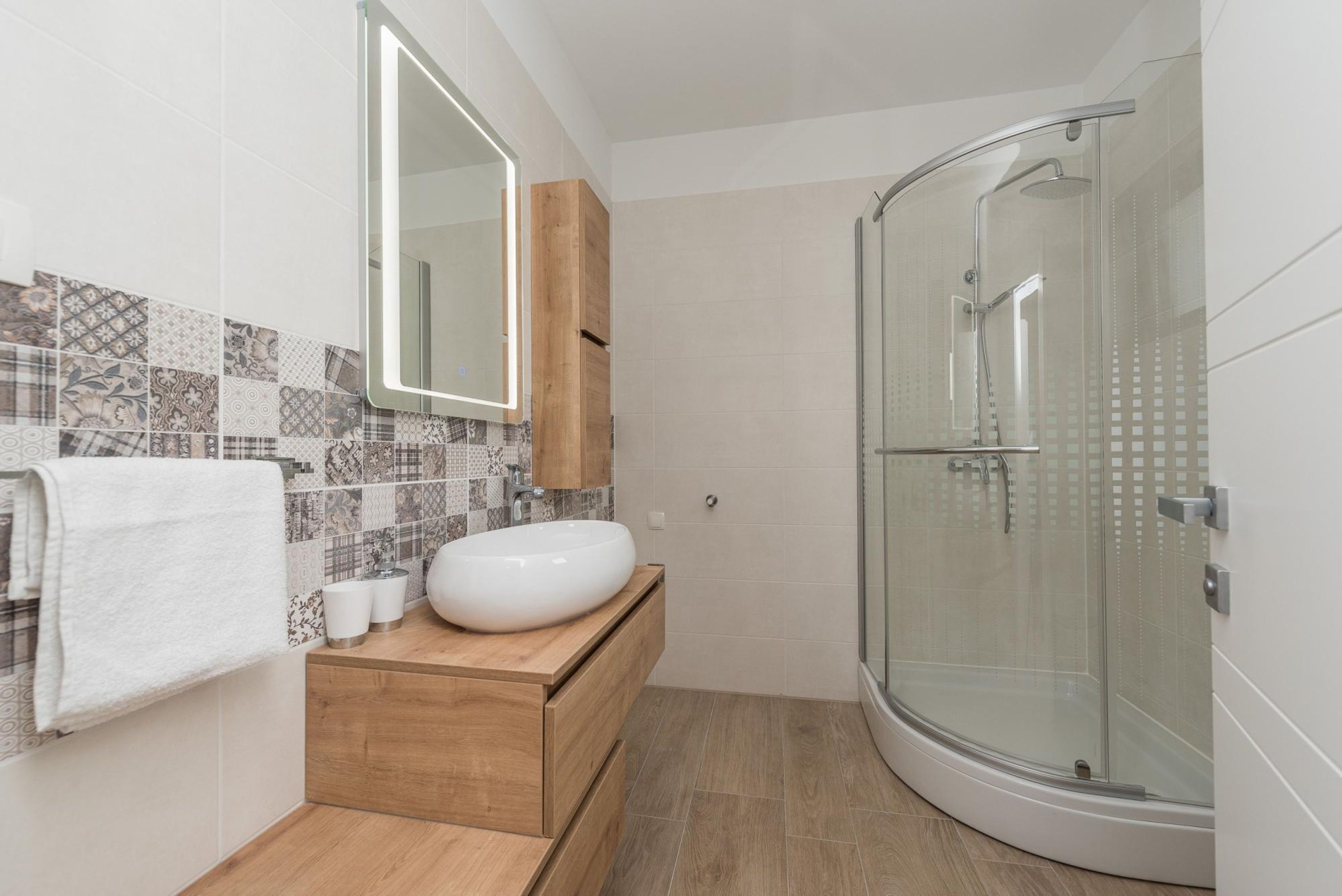 A modern bathroom with a white sink, wooden vanity, patterned tiles, and a glass-enclosed shower.