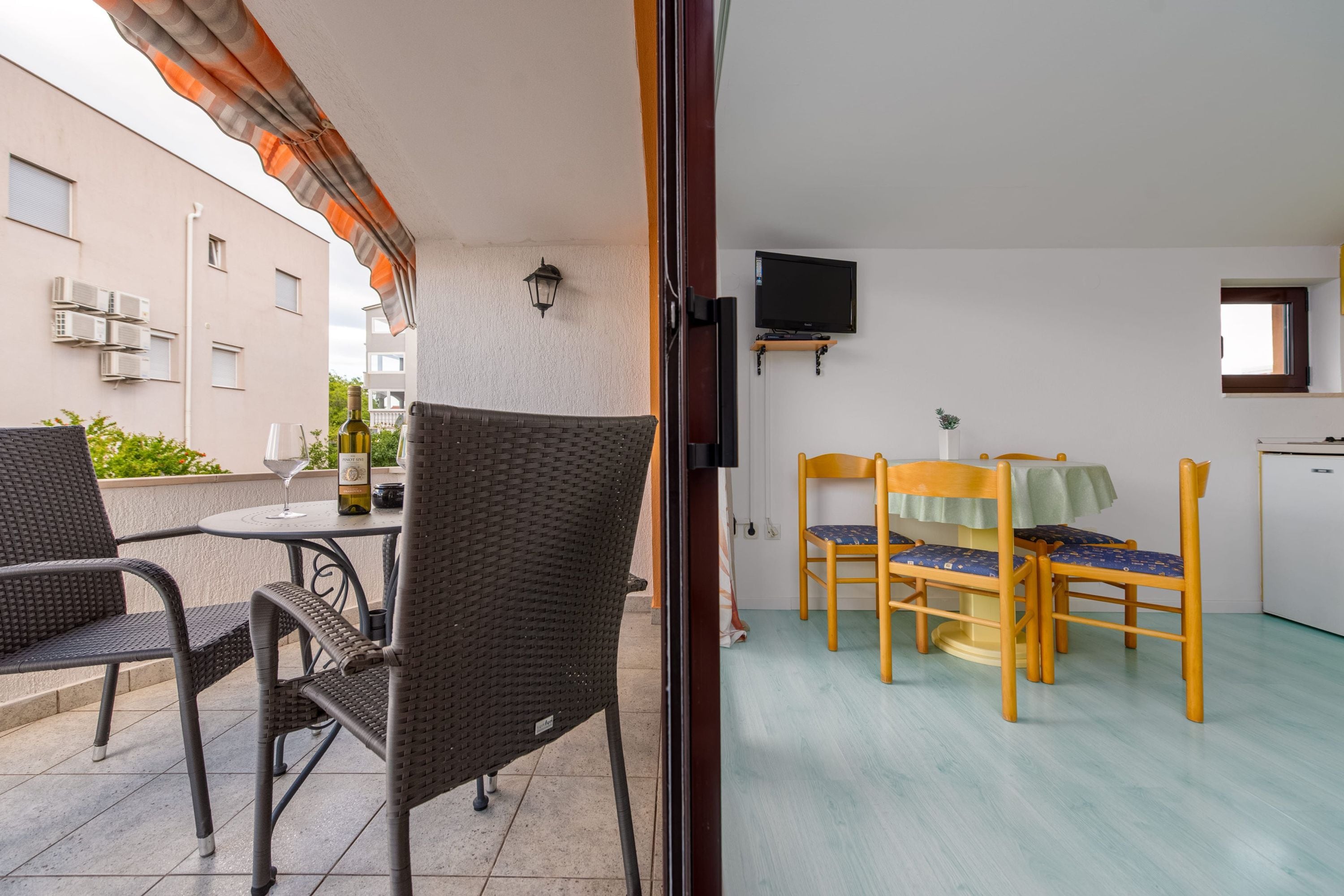 Un balcon avec des fauteuils en osier et une table, une cuisine avec des chaises en bois, et un téléviseur fixé au mur.