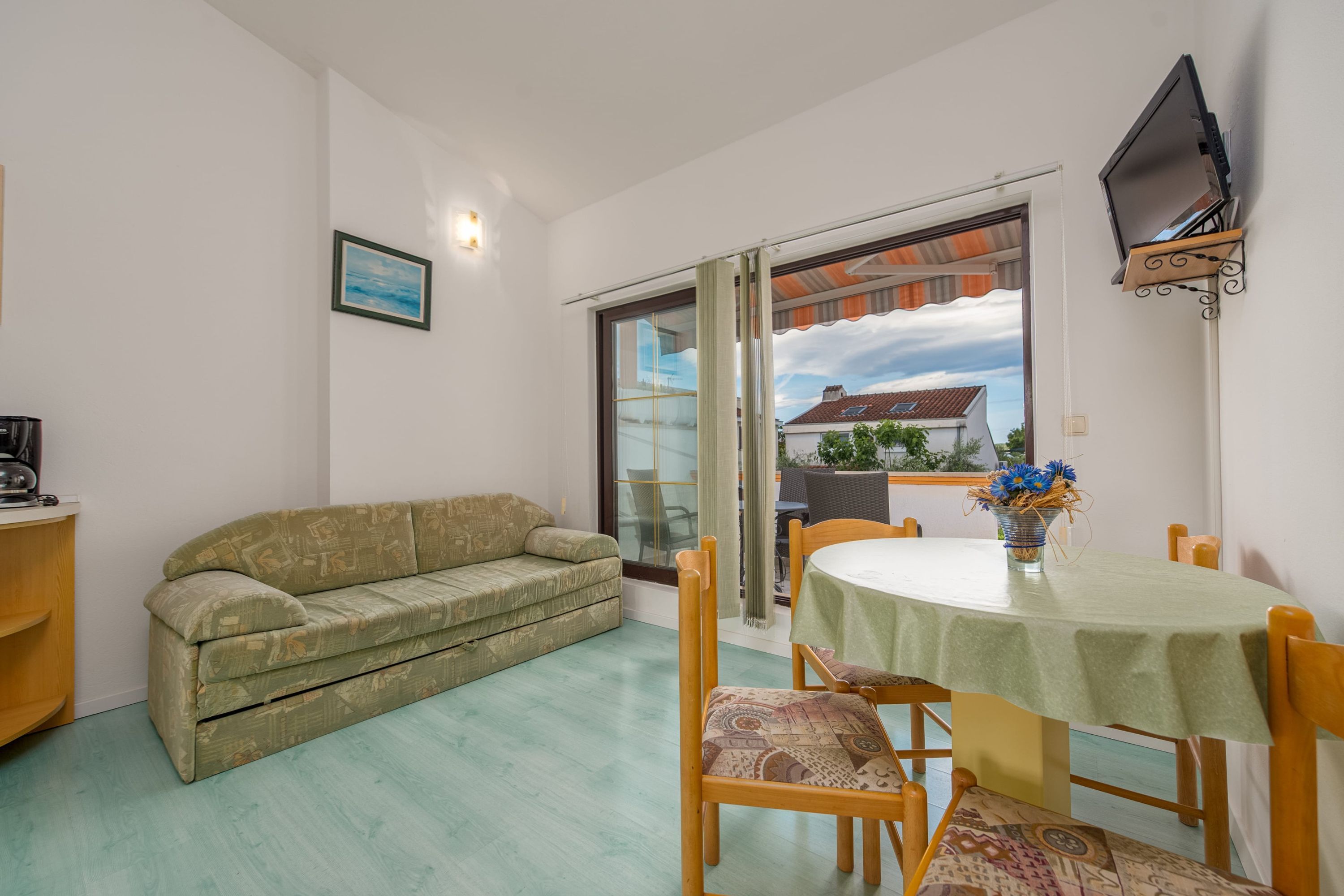 A room with a green sofa, a dining table with chairs, a TV mounted on the wall, and a window with a view of a house.