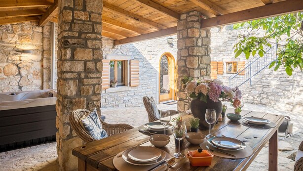 A rustic outdoor dining area with stone walls, wooden beams, and a stone fireplace.