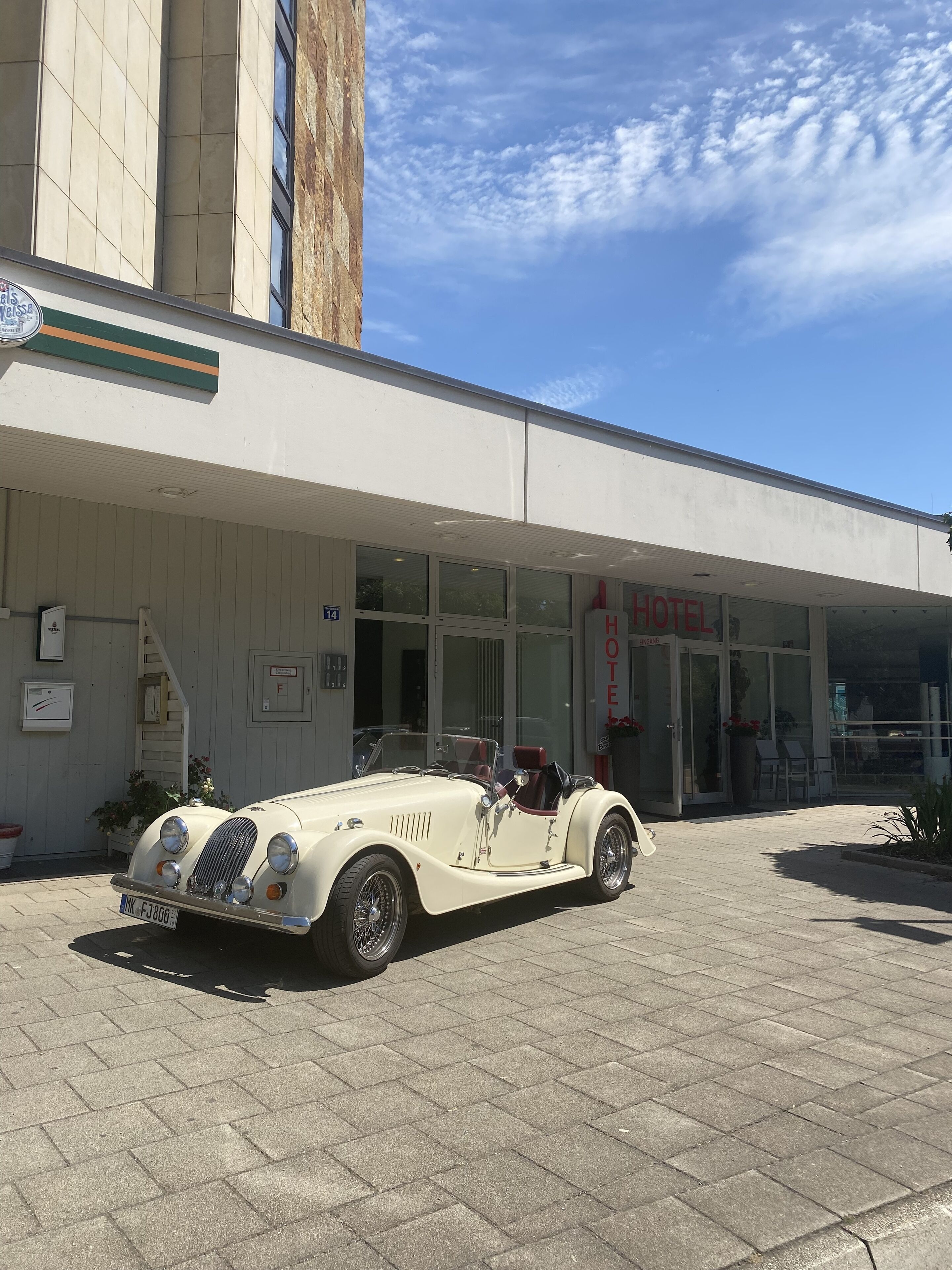 Une voiture décapotable blanche, modèle ancien, est garée devant un hôtel à la façade moderne, sous un ciel dégagé.