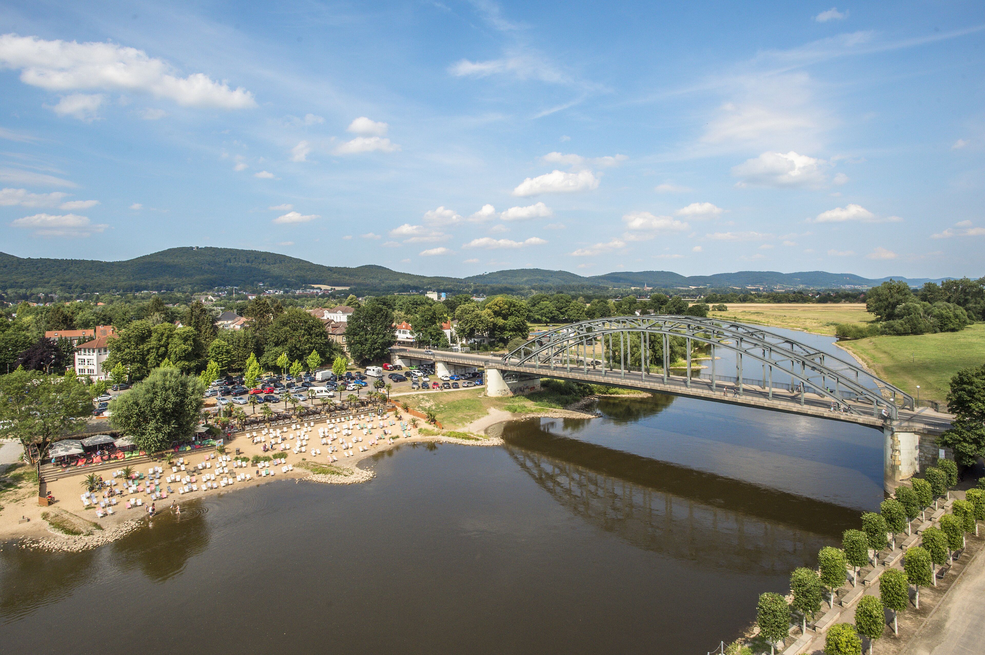 A river with a bridge, a beach with umbrellas, and a town in the background.