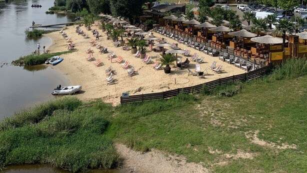 Una playa junto al río con sillones de playa y sombrillas, un barco amarrado y una hilera de edificios al fondo.