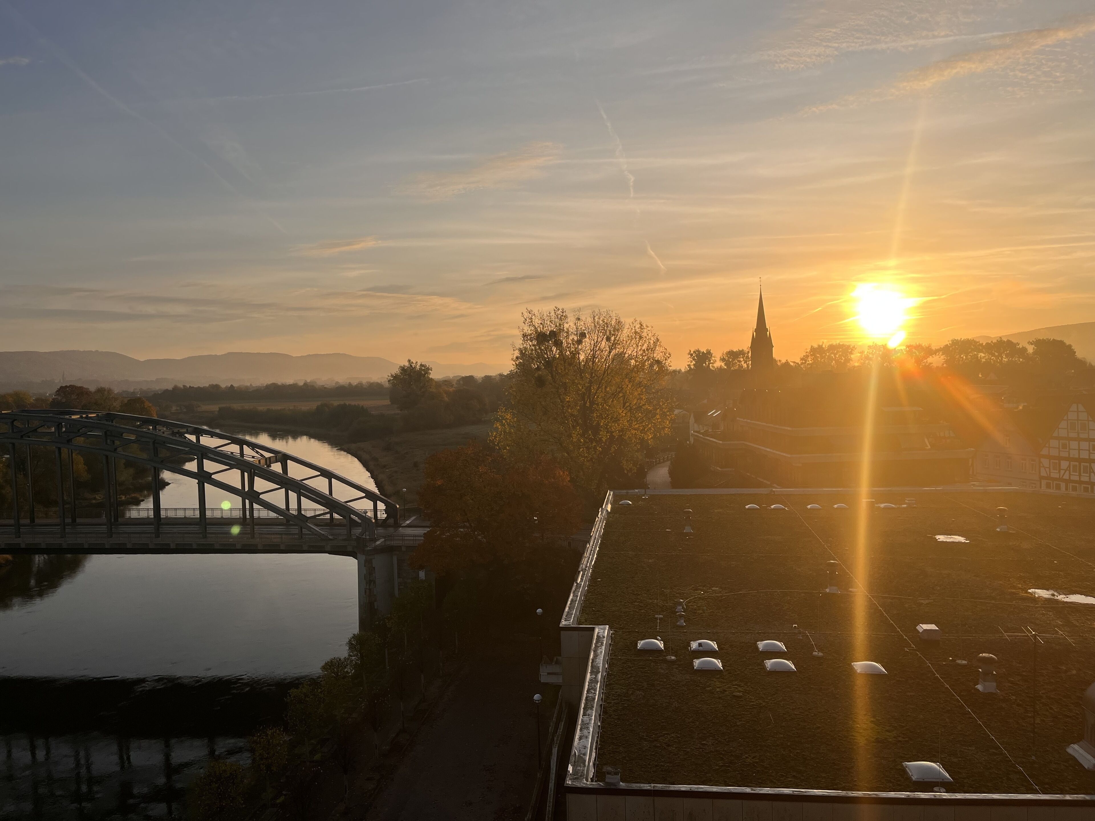 Een brug over een rustige rivier, een kerk met een hoge torenspits, en een veld met verschillende witte gebouwen.