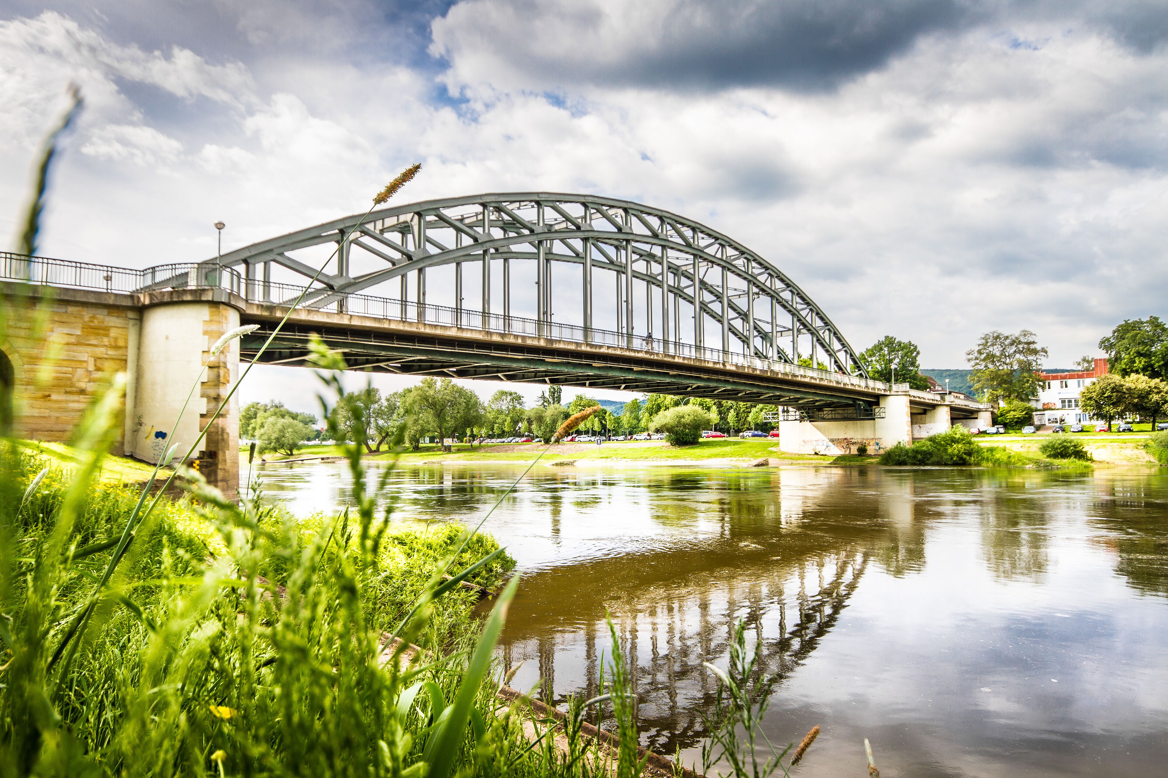 Un pont d’acier enjambe une rivière paisible, bordée de rives verdoyantes et avec des arbres en arrière-plan.