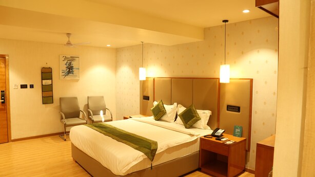 A hotel room with a bed, two chairs, a desk with a phone, and a wall-mounted artwork. - Hotel Reva Regency (Bhopal)