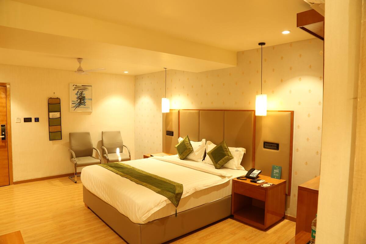 Quarto de hotel com cama, duas cadeiras, escrivaninha com telefone e uma obra de arte na parede.