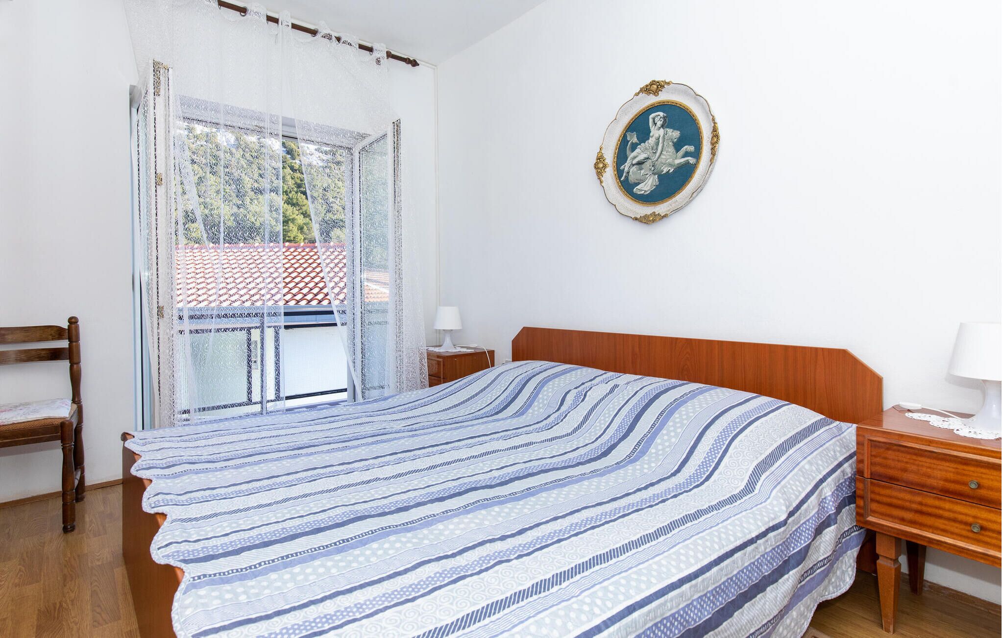 A bedroom with a bed, wooden nightstands, a chair, a window with lace curtains, and a wall-mounted picture frame.