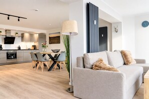 A modern living room with a grey sofa, a dining area with a wooden table and chairs, and a kitchen with built-in appliances.