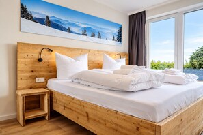 A hotel room with a wooden bed, white bedding, a nightstand, a wall-mounted lamp, and a large window with a view of greenery.