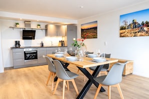 A modern kitchen with built-in appliances, a dining area with a wooden table and chairs, and a painting of a sunset on the wall.